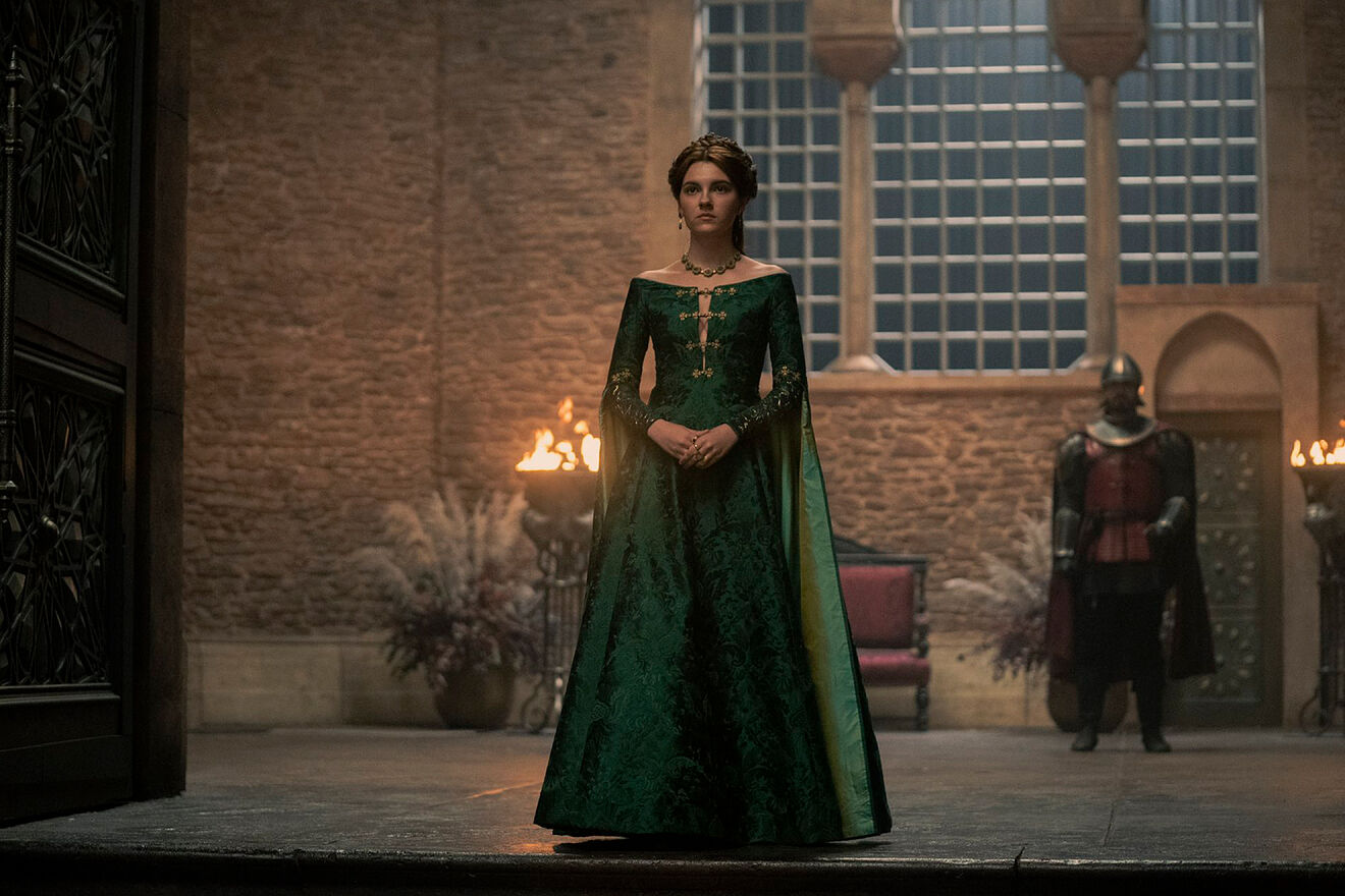 El personaje de Alicent se encuentra en la entrada del salón, de pie, con las manos entrelazadas a la altura de la tripa y vestida con un vestido largo verde